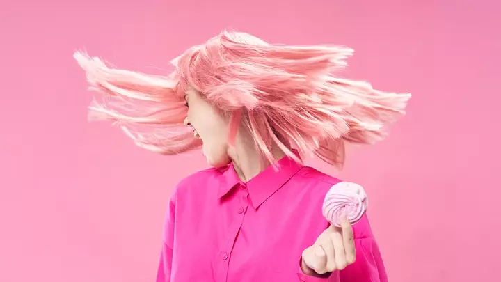 A silver blond-haired woman in pink, on a pink background holding a pink doughnut with hair flying in the air as she turns away from the pastry in her hand
